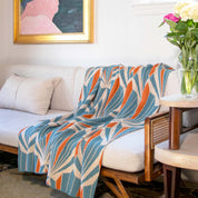 The In2Green Art Deco Shells Throw Blanket, featuring blue, orange, and beige patterns, is draped over a light sofa beside a cushion, a side table with flowers, and a framed painting on the wall.
