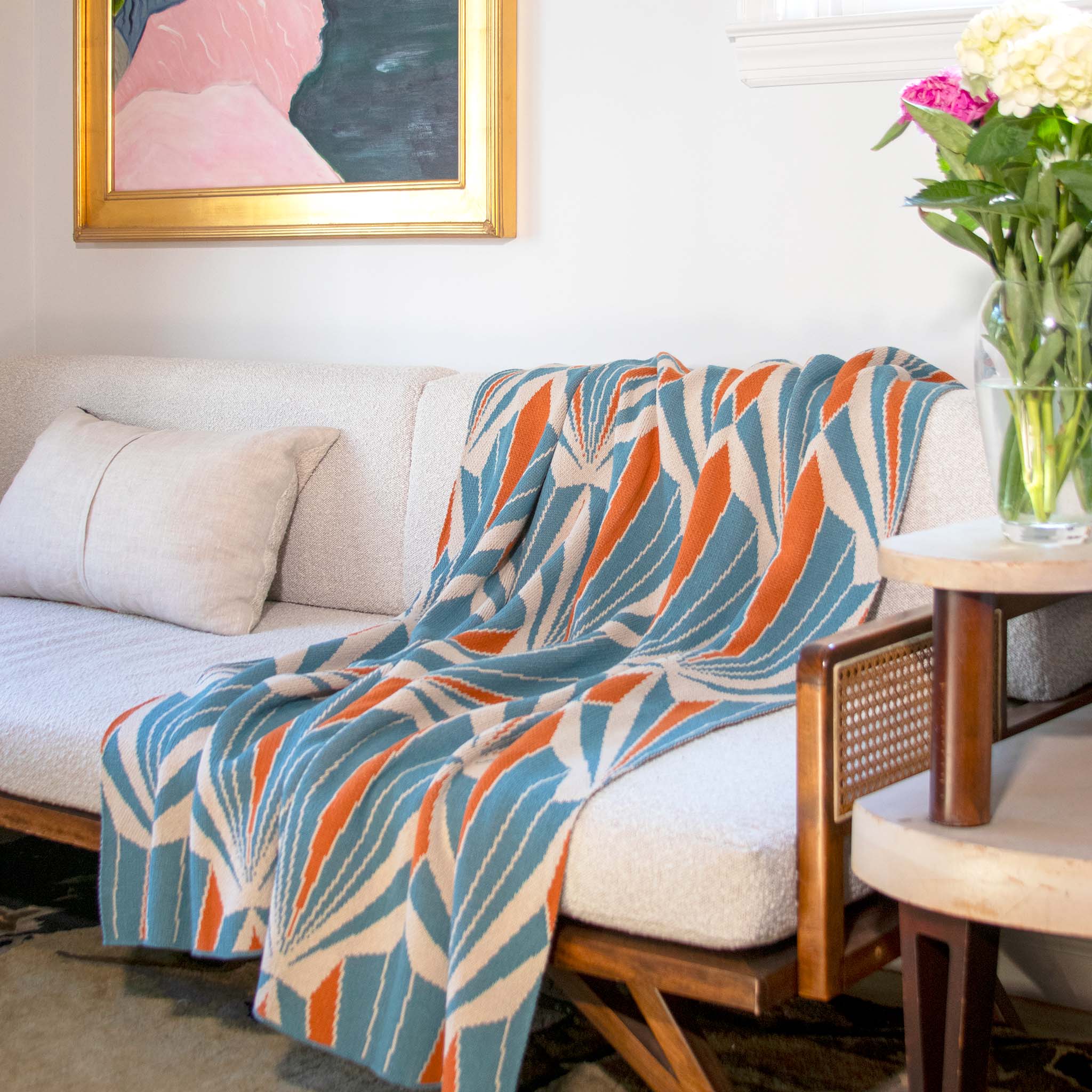 The In2Green Art Deco Shells Throw Blanket, featuring blue, orange, and beige patterns, is draped over a light sofa beside a cushion, a side table with flowers, and a framed painting on the wall.