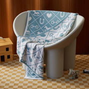 The Baby Pony Quilt Blanket by in2green, featuring blue and white patterns, is draped over a modern white rounded chair on a checkered beige carpet, beside a toy horse and wooden structure.