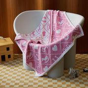 The in2green Baby Pony Quilt Blanket, featuring pink and white heart, anchor, and rocking horse motifs, is draped over a white chair, with a toy horse placed on the nearby checkered floor.