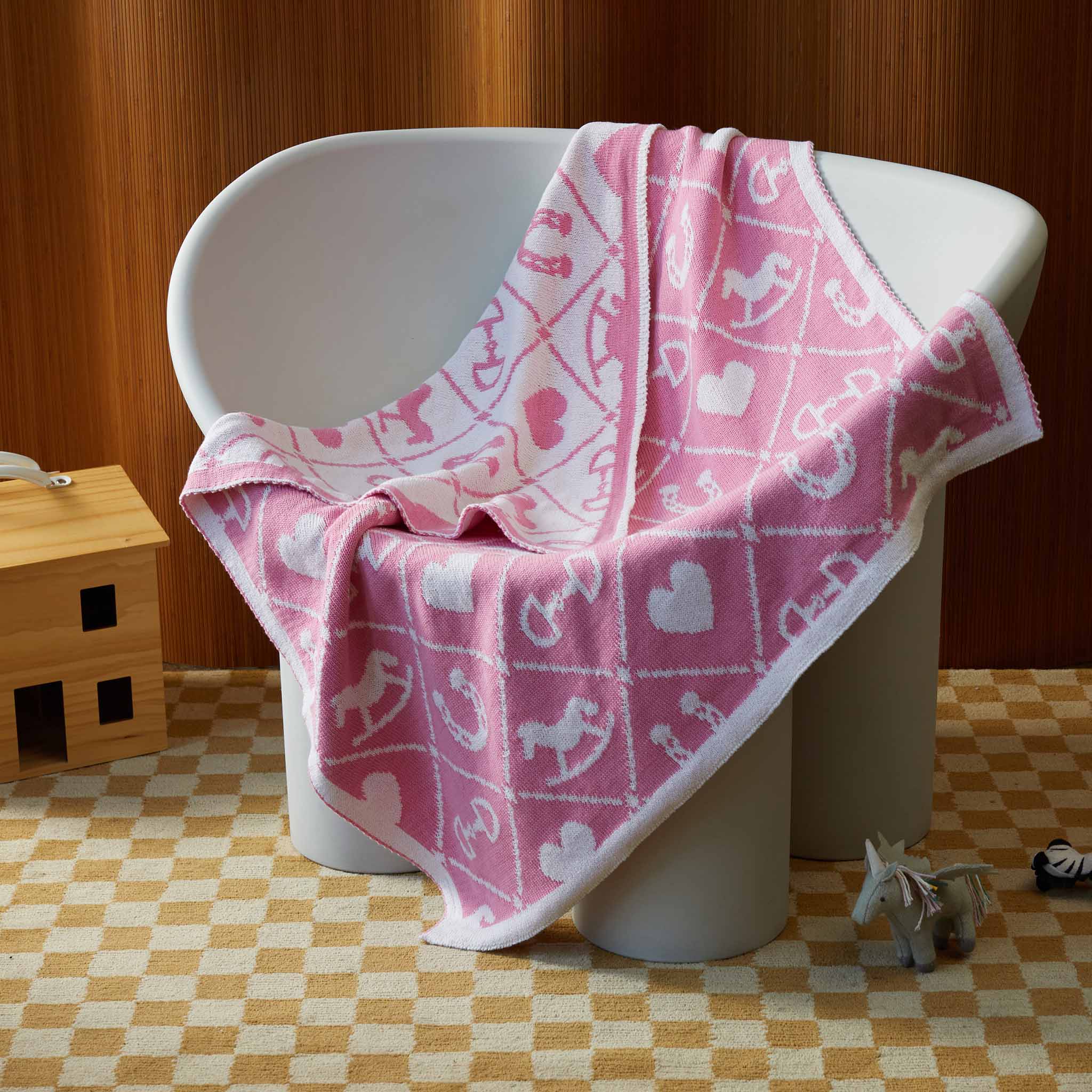 The in2green Baby Pony Quilt Blanket, featuring pink and white heart, anchor, and rocking horse motifs, is draped over a white chair, with a toy horse placed on the nearby checkered floor.