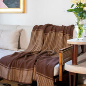 The in2green Block Canyon Throw Blanket is draped over a light sofa, next to a wooden chair and a vase of flowers on a side table.