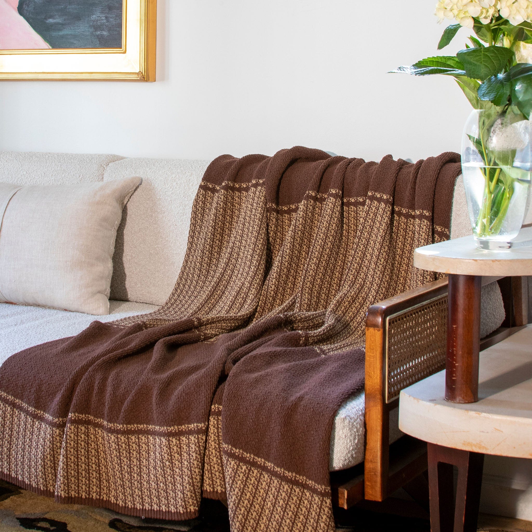 The in2green Block Canyon Throw Blanket is draped over a light sofa, next to a wooden chair and a vase of flowers on a side table.