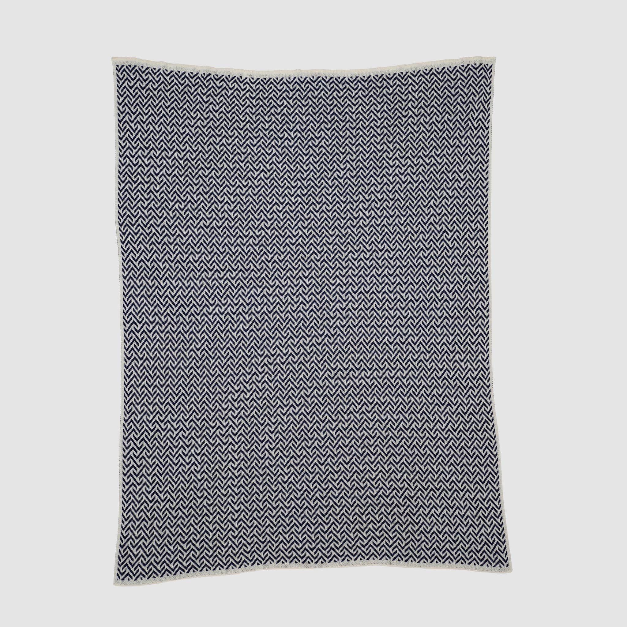 The Broken Zig Zag Throw Blanket showcases a repeating black and white chevron pattern, crafted from recycled cotton yarns for a sustainable addition to modern decor.
