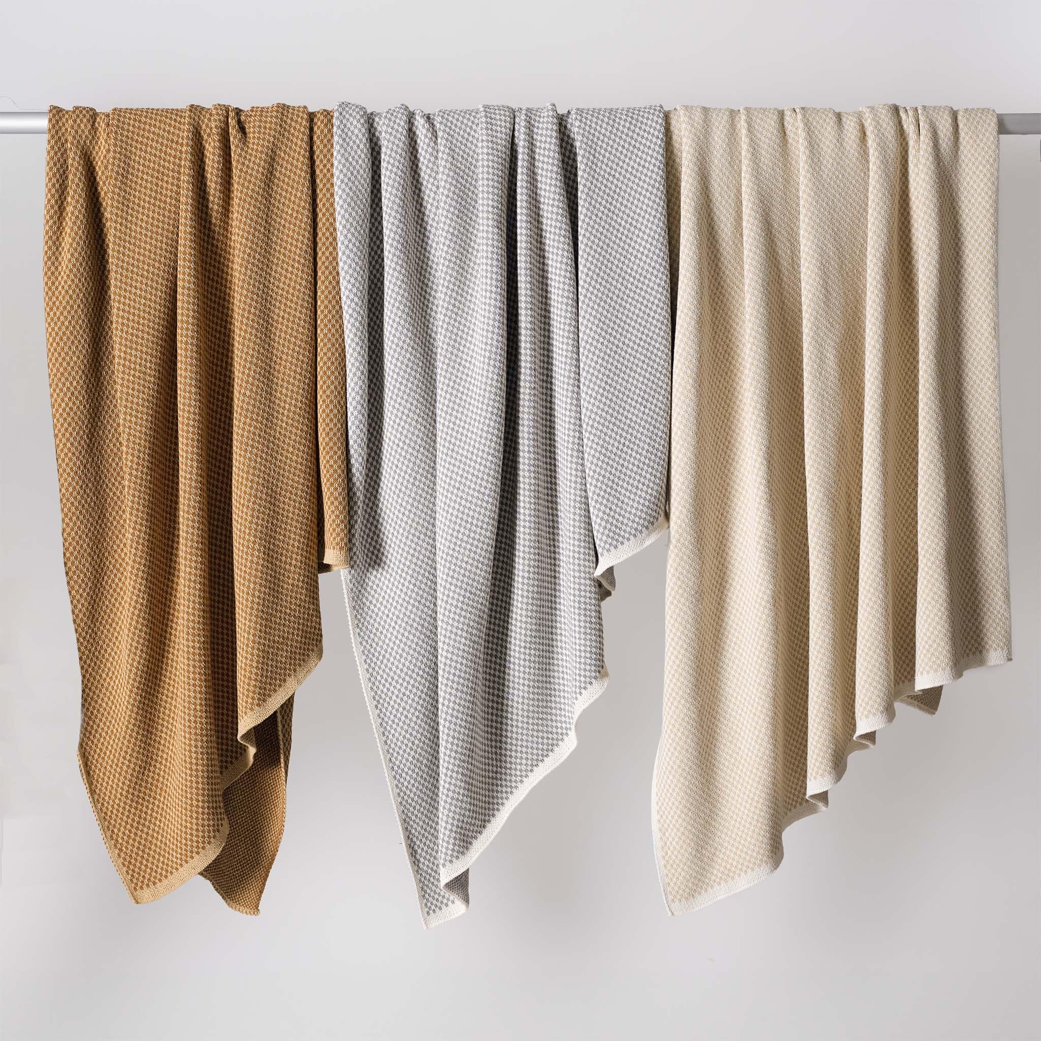 Three in2green Checkerboard Throw Blankets in brown, gray, and cream hang over a horizontal rod against a plain white background.
