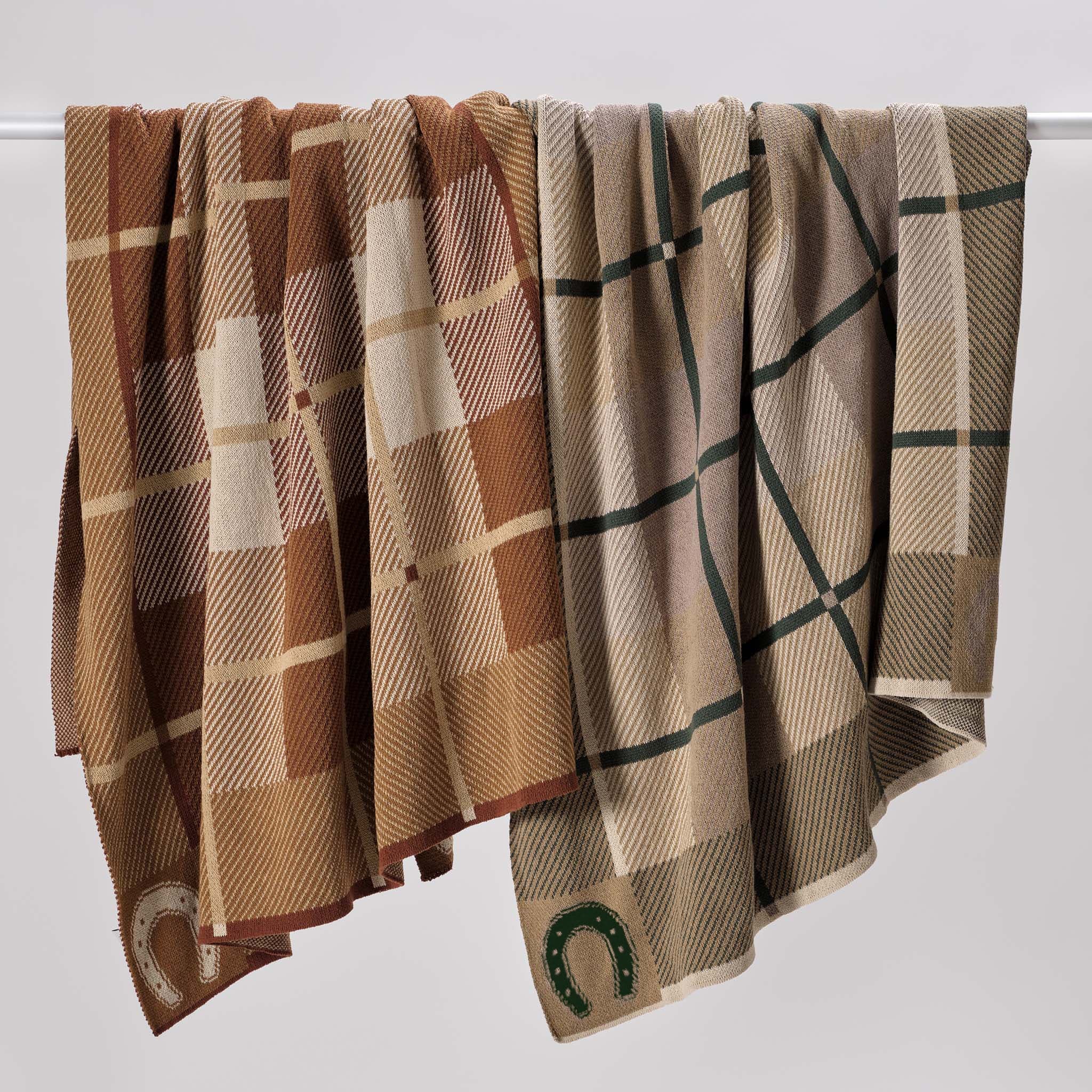 Three In2Green Equestrian Barn Plaid Throw Blankets in brown and beige, made from upcycled cotton and featuring embroidered horseshoe motifs, are draped over a horizontal bar against a plain background.