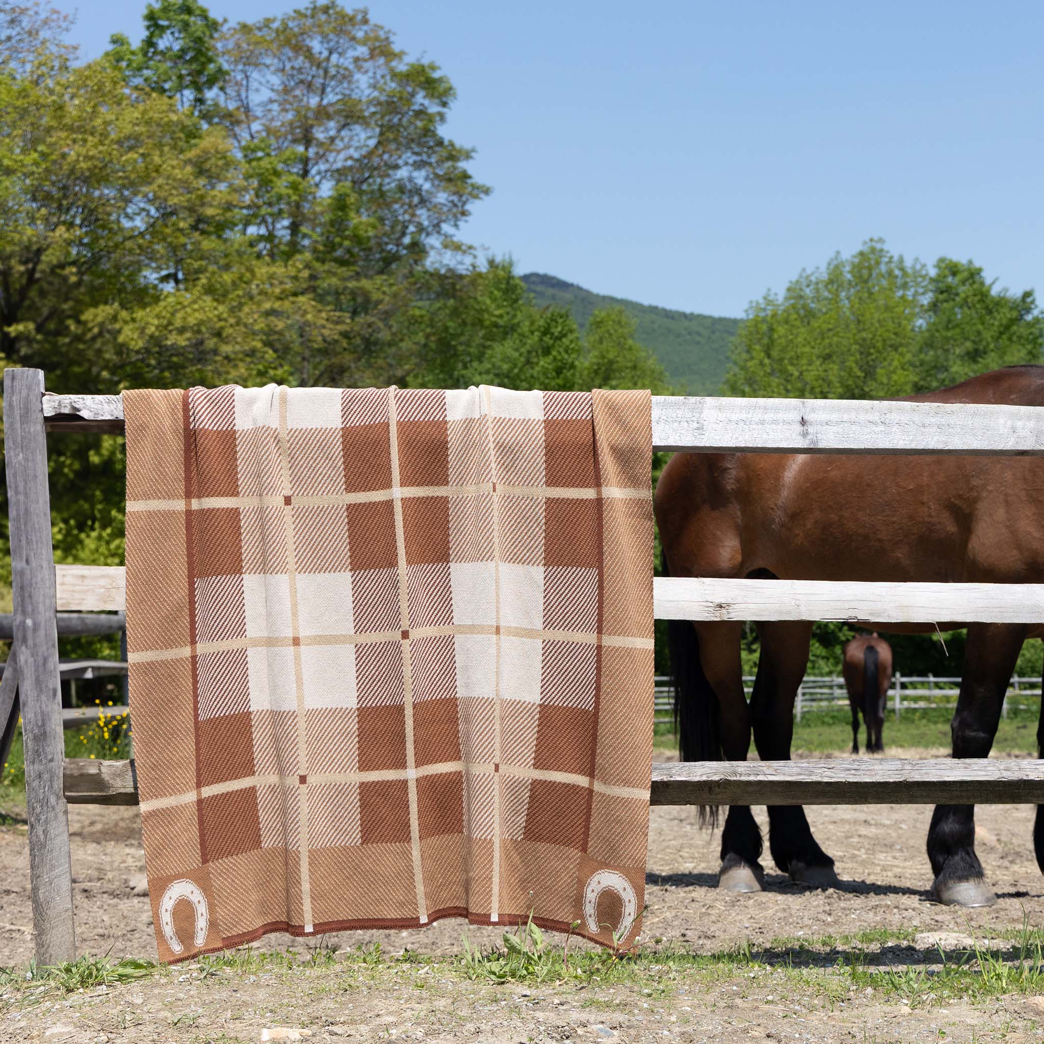 The In2Green Equestrian Barn Plaid Throw Blanket, made from upcycled cotton and recycled polyester and featuring horseshoe designs, is draped over a wooden fence beside a horse in a rustic outdoor setting.