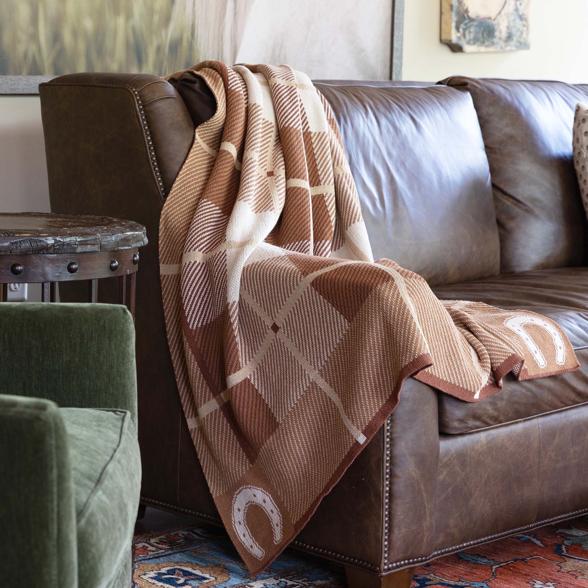 The In2Green Equestrian Barn Plaid Throw Blanket, made from recycled polyester, showcases a brown and beige check with horseshoe designs, elegantly displayed over a brown leather sofa in the living room.