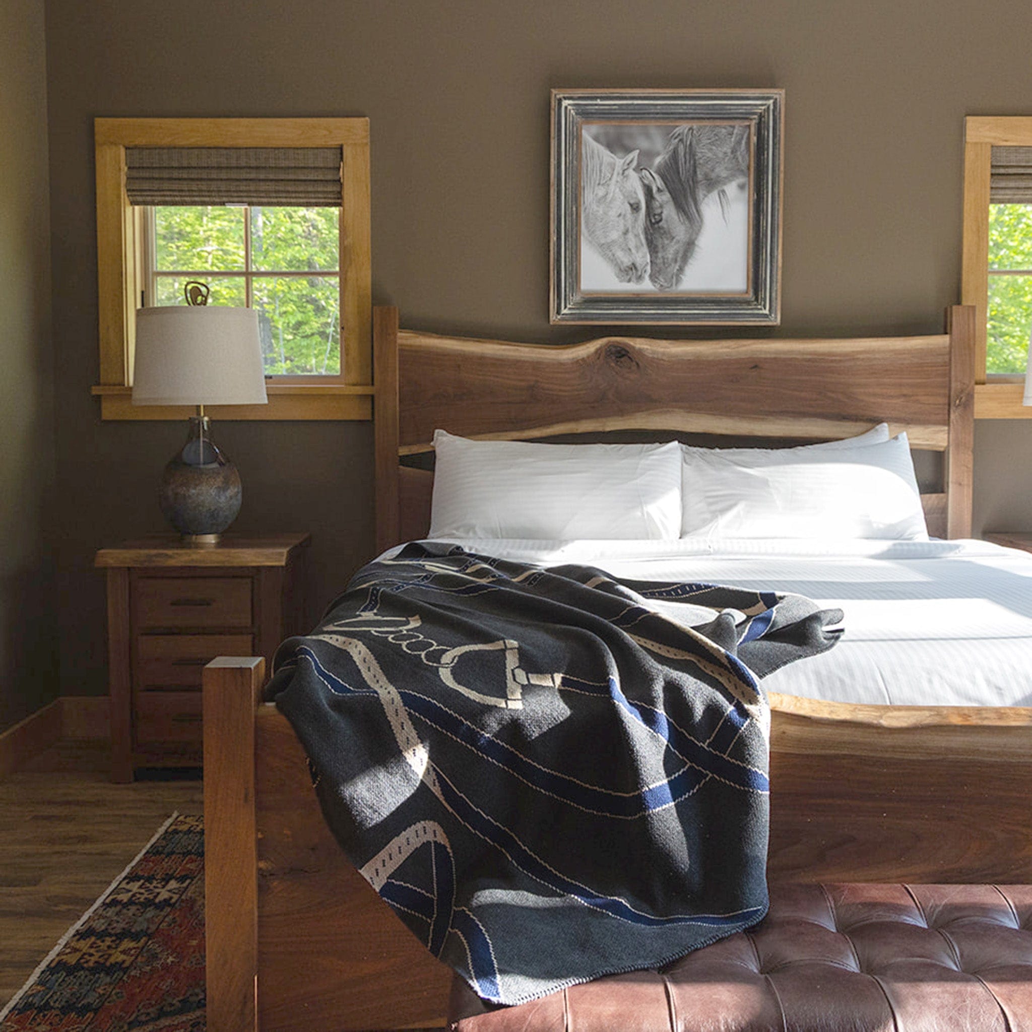 A rustic bedroom features a neatly made bed with white linens and the In2Green Equestrian Tack Throw Blanket, wooden furniture, and a horse painting above—perfect for horse lovers.
