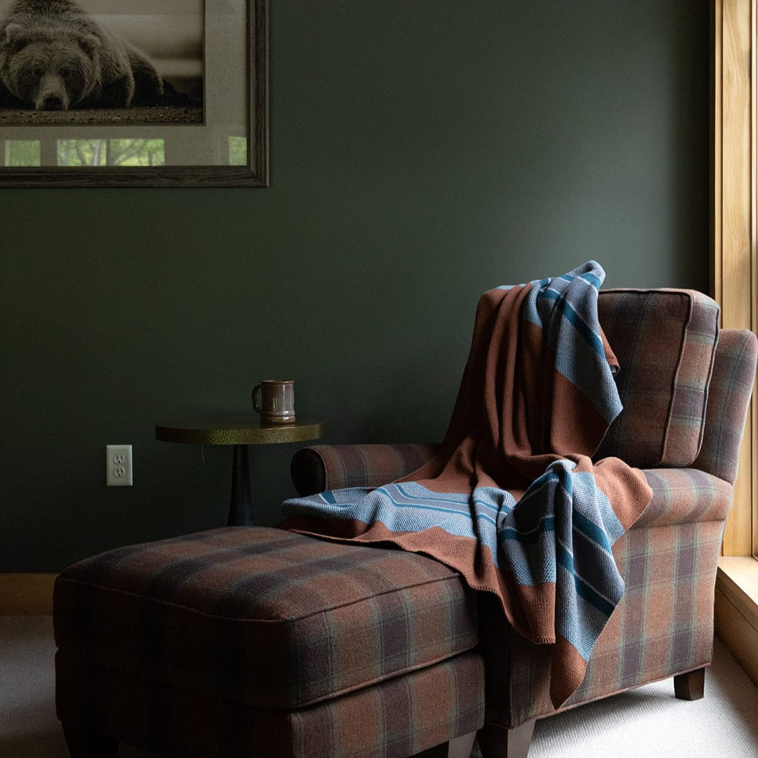 A plaid armchair with ottoman sits by a window, draped with a blue and brown Lodge Stripe Throw Blanket. A side table holds a ceramic mug, while a framed photo of a bear hangs on the dark green wall.