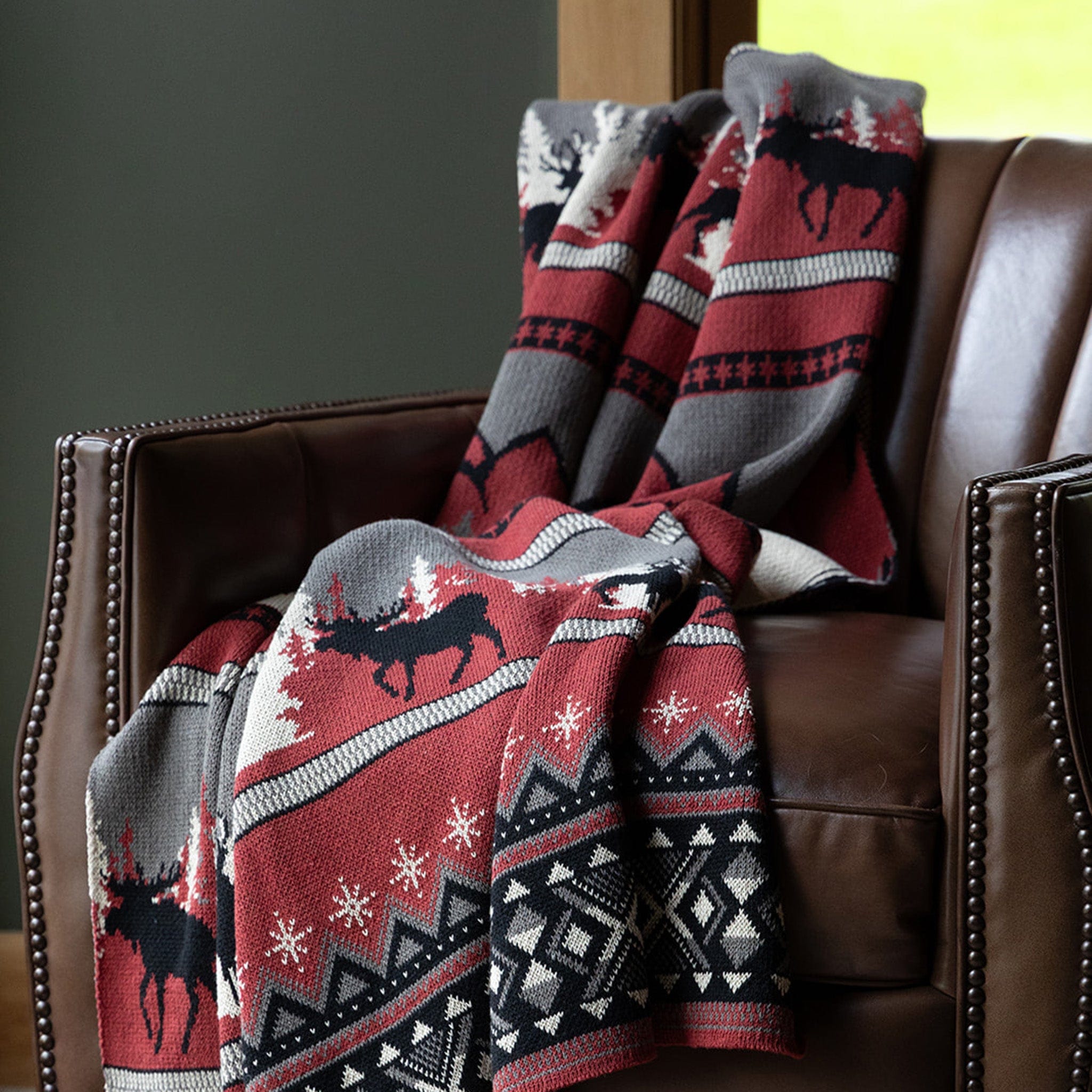 The Mountain Fair Isle Throw Blanket, featuring red, black, white, and gray moose and geometric patterns in recycled cotton, adds cozy holiday style when draped over a brown leather armchair.