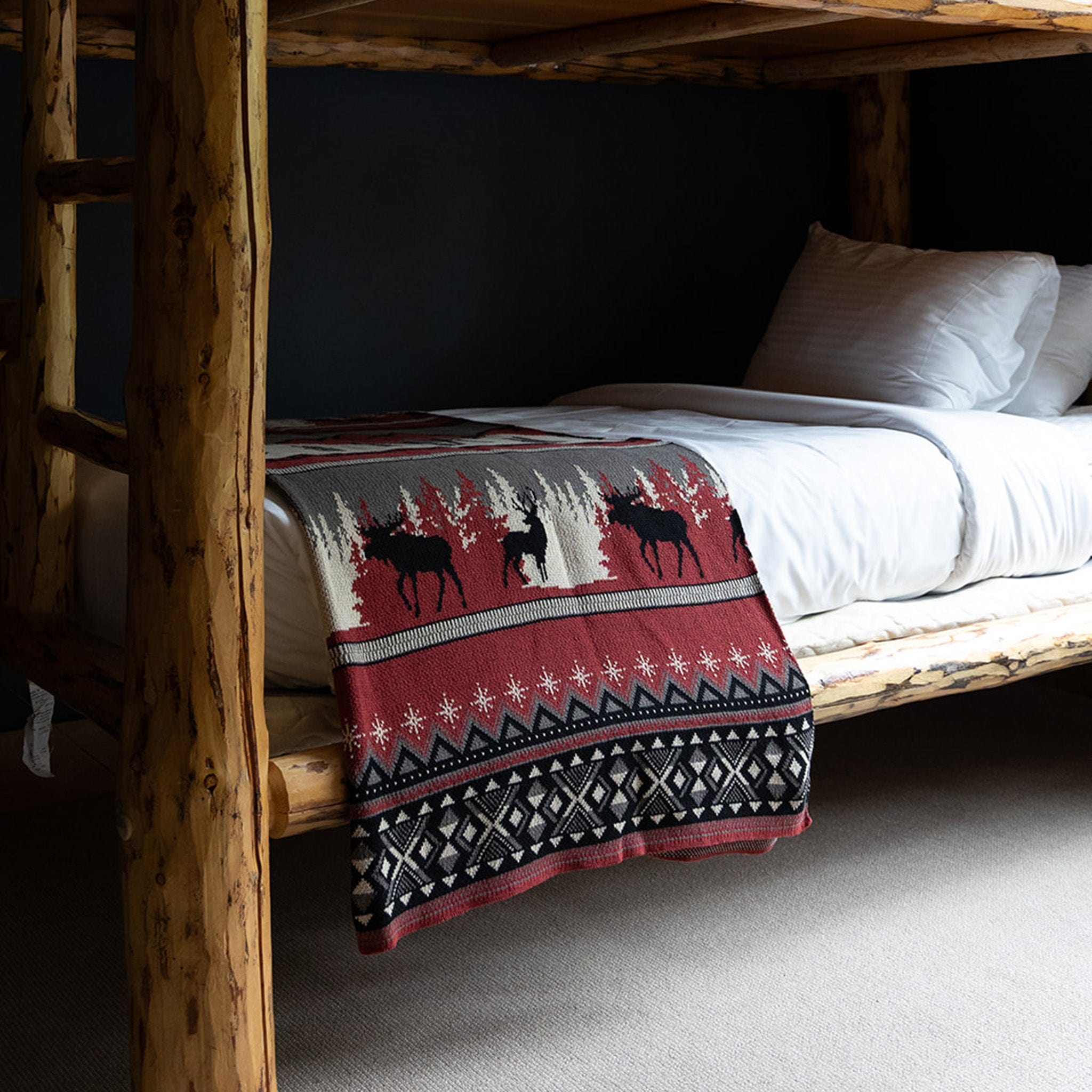 A log-frame bunk bed with white bedding is styled for the holidays with a Mountain Fair Isle Throw Blanket—recycled cotton, red, black, and white moose and tree patterns—draped over the lower bunk for a cozy festive touch.