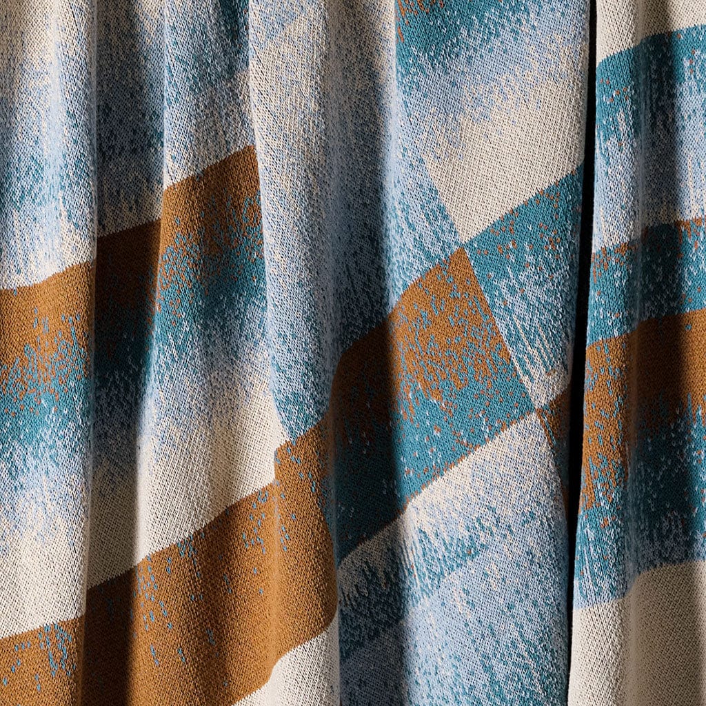 Close-up of the in2green Plaid Ombre Throw Blanket, featuring a modern geometric plaid pattern with diagonal blue, brown, and off-white stripes. Textured material drapes in folds, adding stylish depth to this cozy accent.