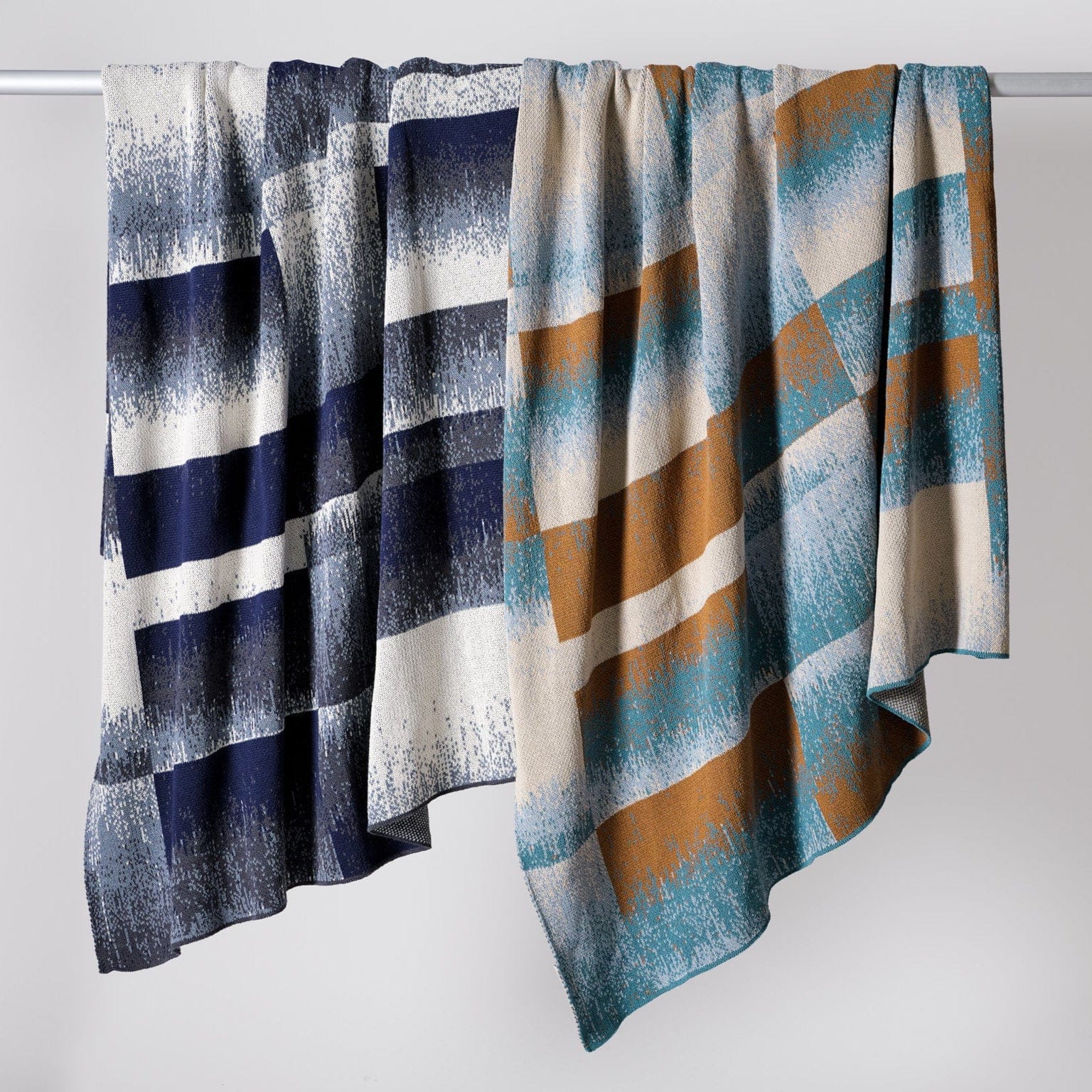 Two patterned blankets hang side by side on a rod, including the Plaid Ombre Throw Blanket, which features a classic check design in blue, white, teal, and mustard tones. Made with recycled yarn for an eco-friendly touch.
