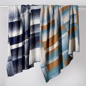 Two patterned blankets hang side by side on a rod, including the Plaid Ombre Throw Blanket, which features a classic check design in blue, white, teal, and mustard tones. Made with recycled yarn for an eco-friendly touch.