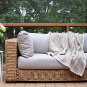 An in2green Poly Herringbone Edge Throw Blanket made from recycled polyester rests on a wicker sofa with light gray cushions on a wooden deck, with trees in the background.