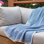The in2green Poly Herringbone Edge Throw Blanket in blue and white is draped over a light gray outdoor sofa with a wicker base, placed by a window with lush greenery outside.