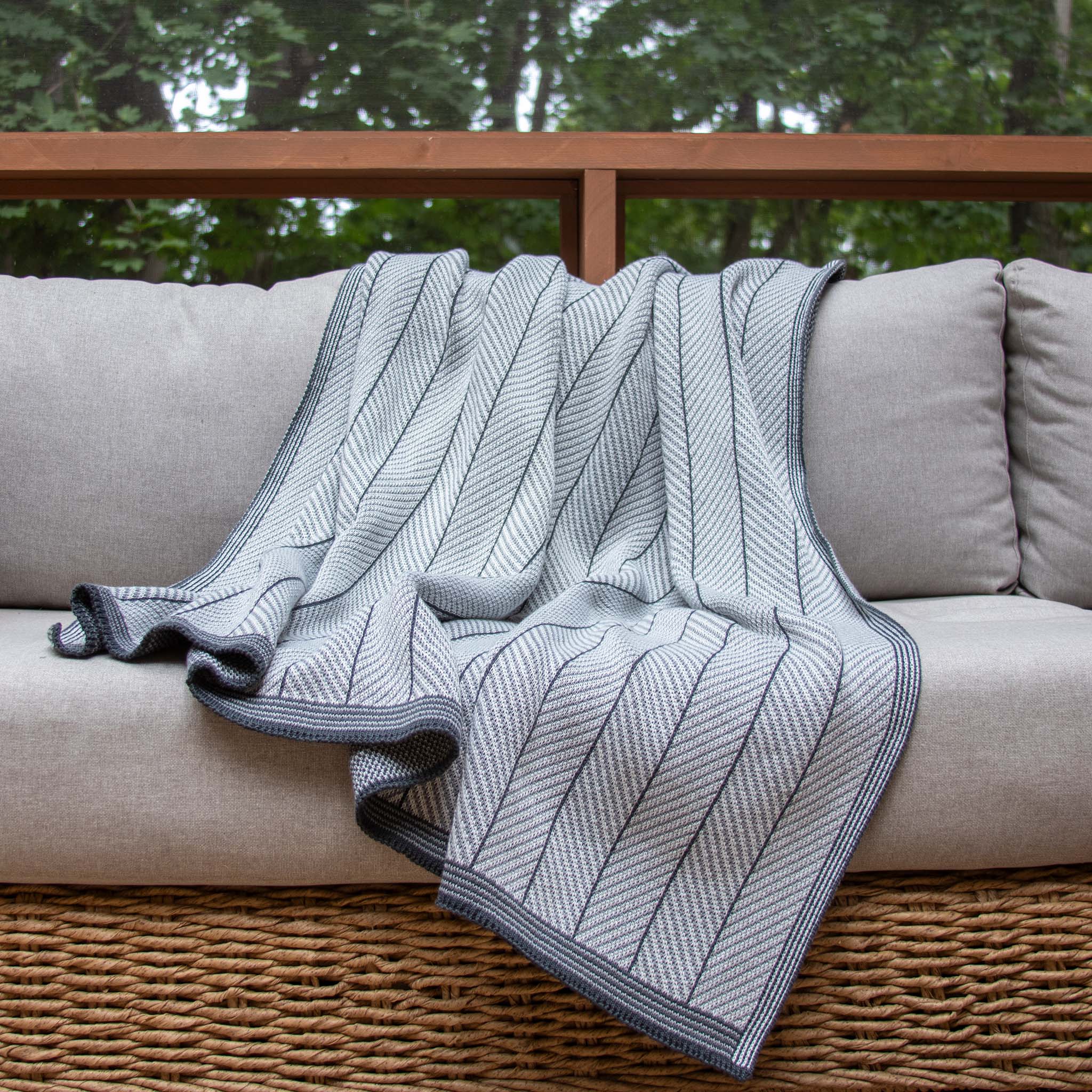A Poly Herringbone Edge Throw Blanket by in2green in light gray is draped over a light beige cushioned wicker sofa, placed before a window with a view of green trees.