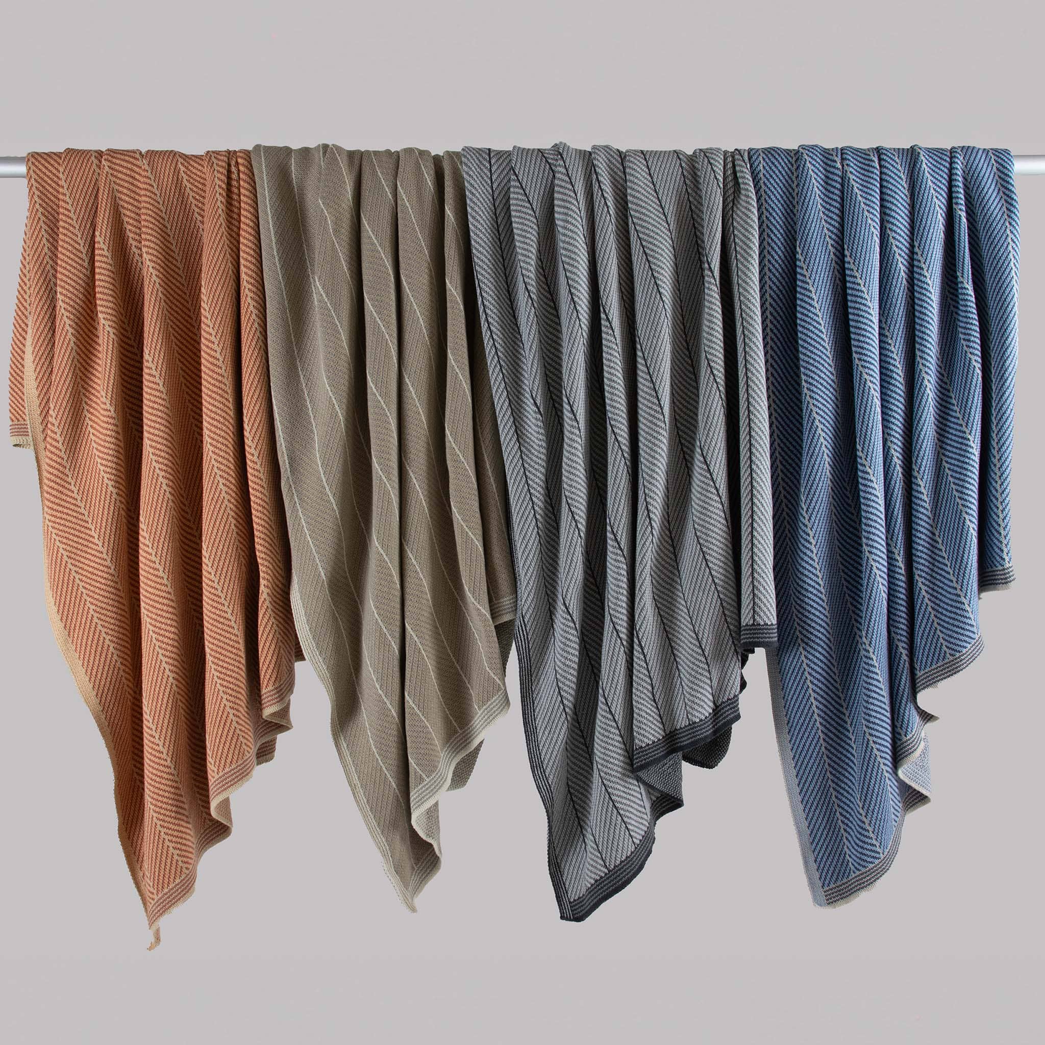 Four in2green Poly Herringbone Edge Throw Blankets in orange, beige, gray, and blue hang neatly on a rod, displaying their distinctive herringbone knit against a plain background.