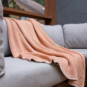 A Poly Herringbone Edge Throw Blanket by in2green, in peach and made from recycled polyester, is draped over a light gray cushioned sofa near a window.