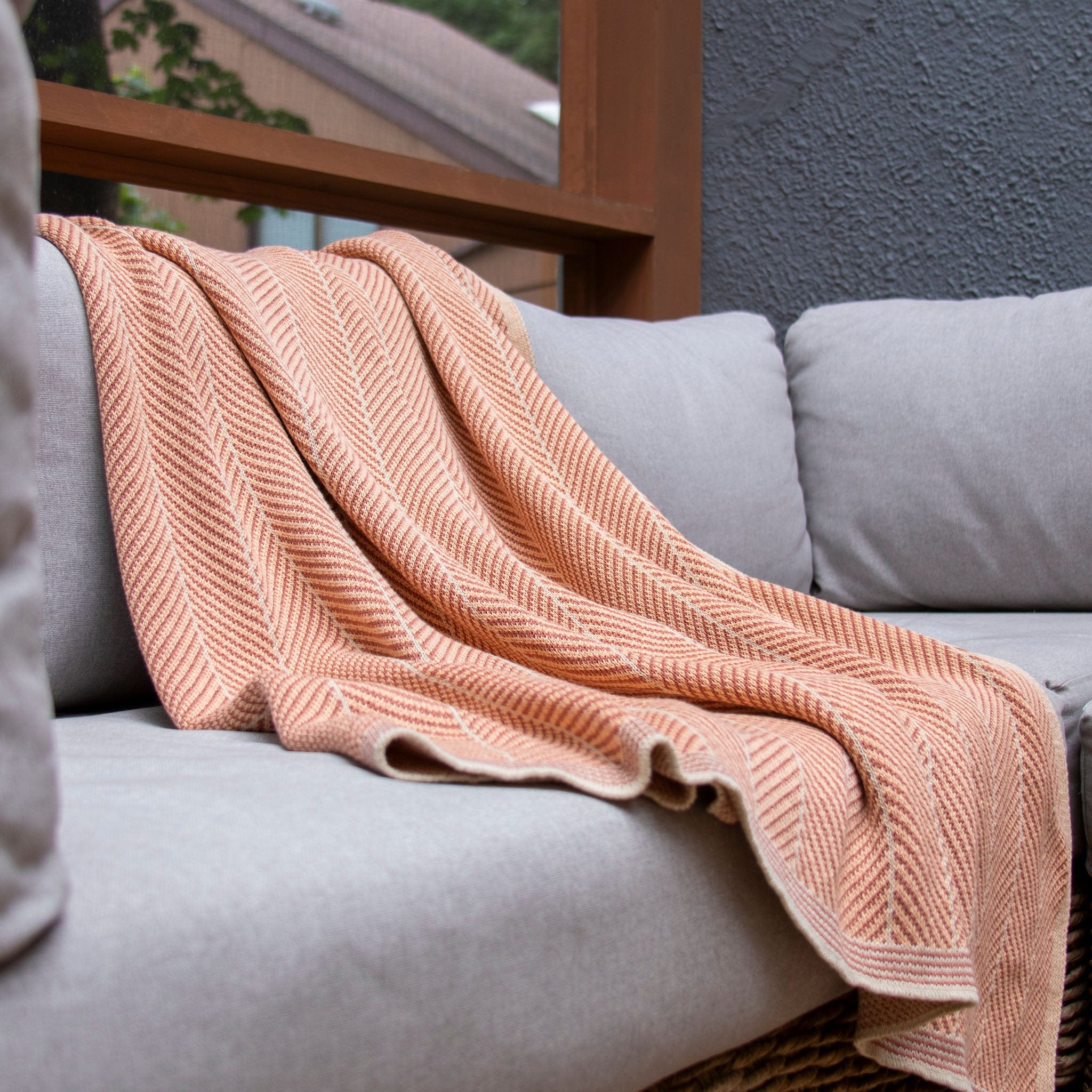 A Poly Herringbone Edge Throw Blanket by in2green, in peach and made from recycled polyester, is draped over a light gray cushioned sofa near a window.