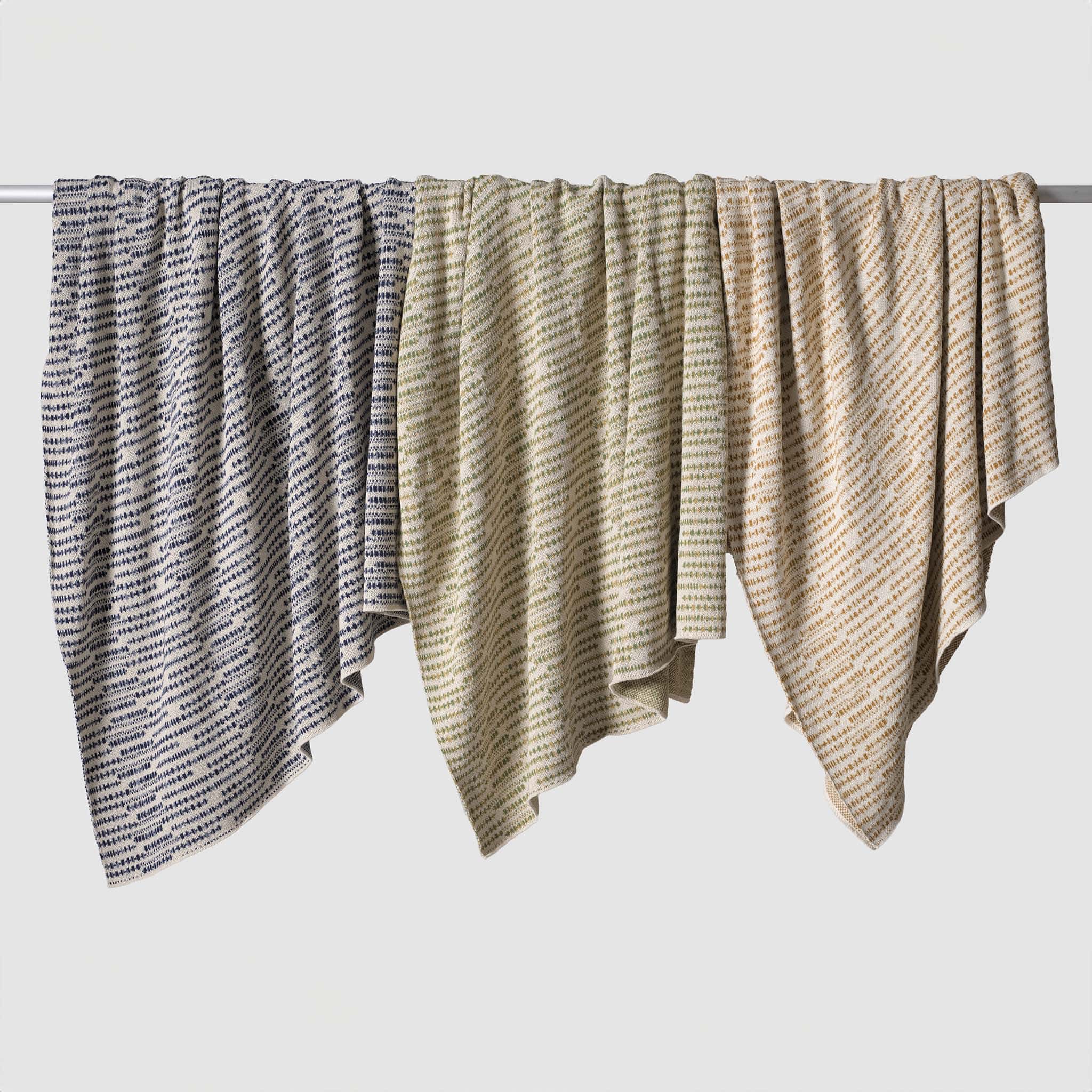 Three In2Green Sand Stone Throw Blankets, made with eco-friendly yarns, are draped over a rod against a white background, bringing organic charm to any space.