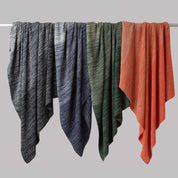 Four in2green Textured Ombre Throw Blankets in black, navy, green, and orange hang side by side on a horizontal rod against a plain gray background.