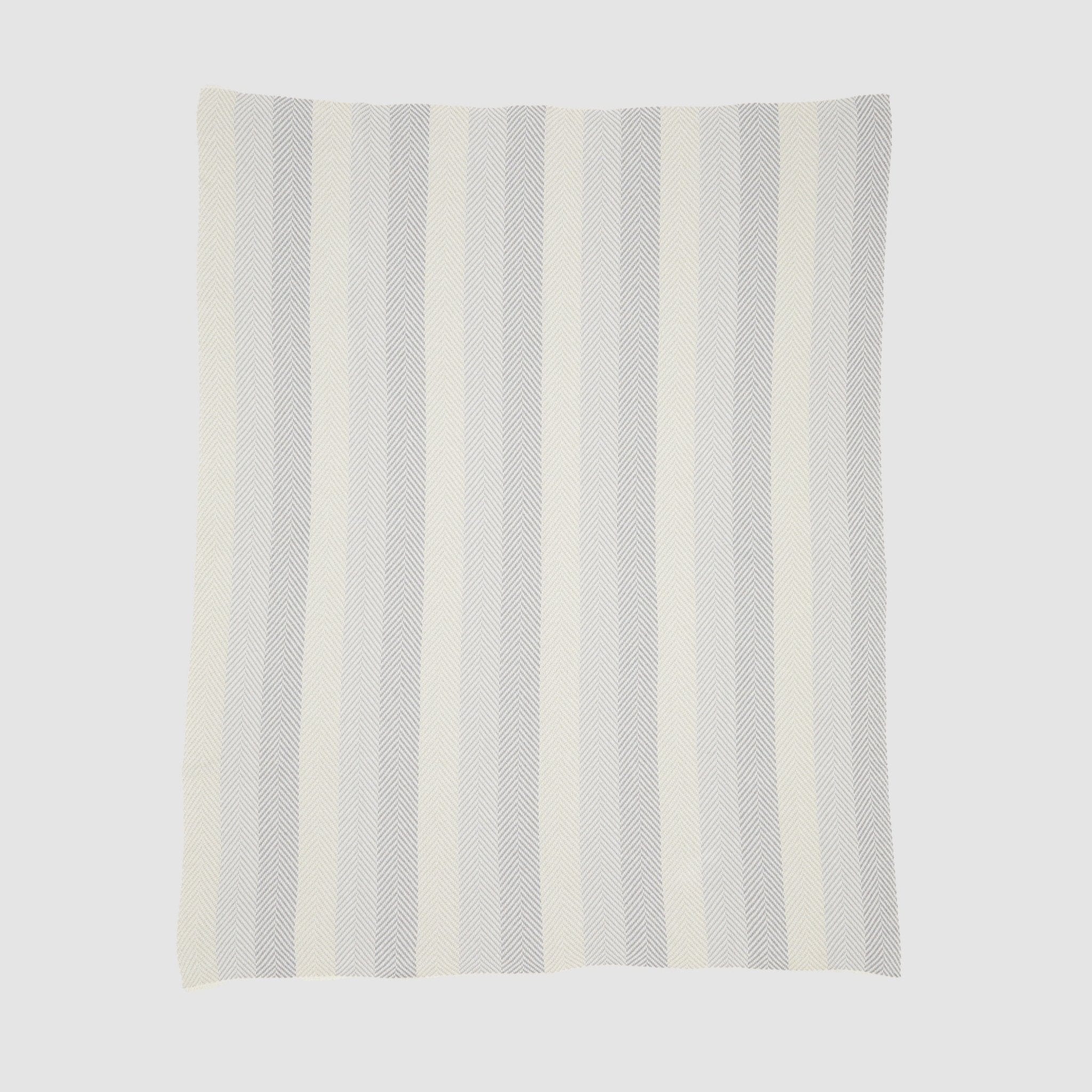 The Herringbone Hues Throw Blanket from In2Green, made in the USA with recycled cotton, features a rectangular design with alternating vertical stripes in light gray and off-white against a plain background.