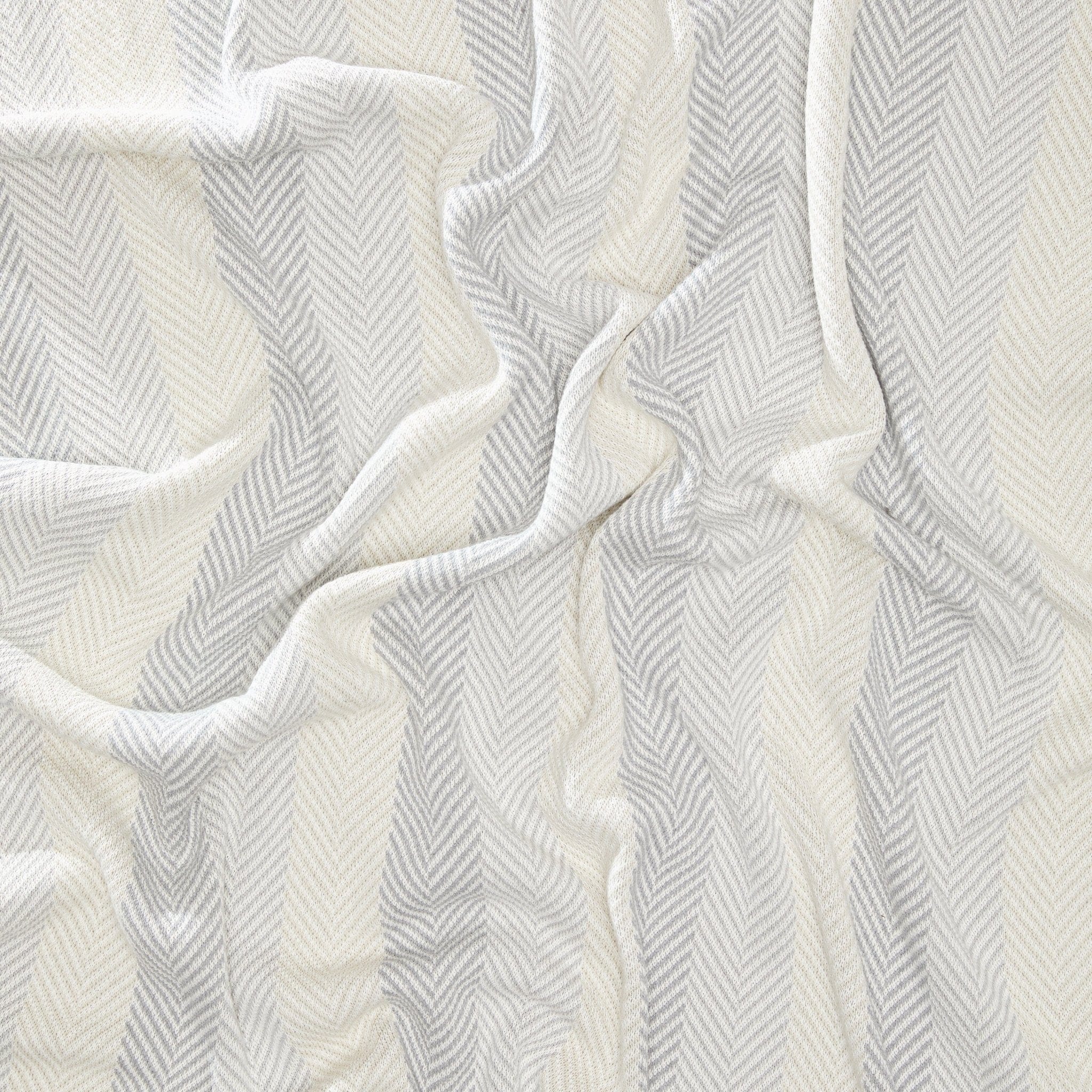 The Herringbone Hues Throw Blanket by In2Green is crafted in the USA from recycled cotton, featuring a wavy drape with a chevron pattern in alternating shades of cream and light gray.