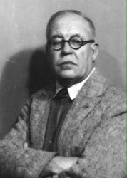 A person with short hair and round glasses stands with crossed arms, wearing a textured jacket decorated with the Pavot Throw Blanket by Maurice Pillard Verneuil from in2green, introducing an Art Nouveau flair to the contemporary scene.