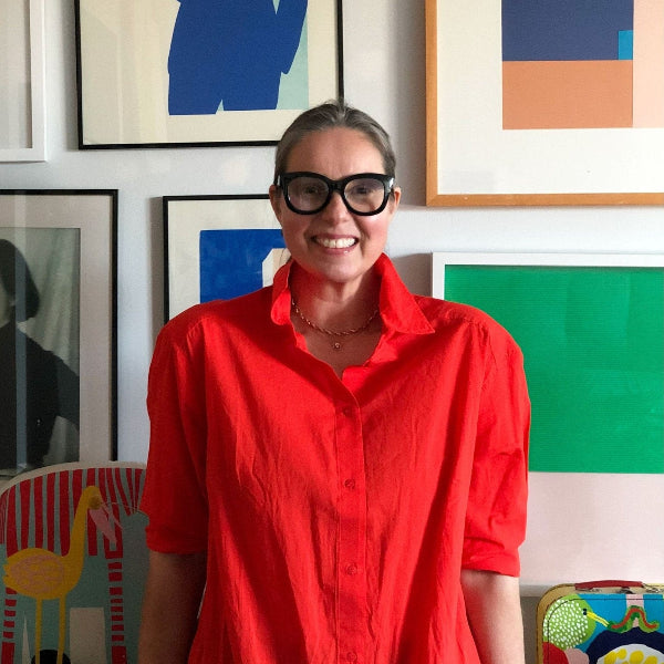 A person in a red shirt and black glasses smiles in front of a wall featuring framed abstract art and the vibrant Poly Kaareva Throw Blanket by Petra Kaksonen from in2green, adding eco-friendly charm.