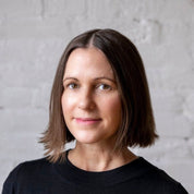 A person with shoulder-length brown hair wearing a black top stands against a white brick wall, reminiscent of an urban landscape, similar to the modern aesthetic of the Level Throw Blanket by Kelly Harris Smith from in2green.