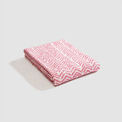 A folded Warrior Throw Blanket Pink by In2Green, featuring a pink and white chevron pattern, rests on a gray surface, symbolizing breast cancer support.