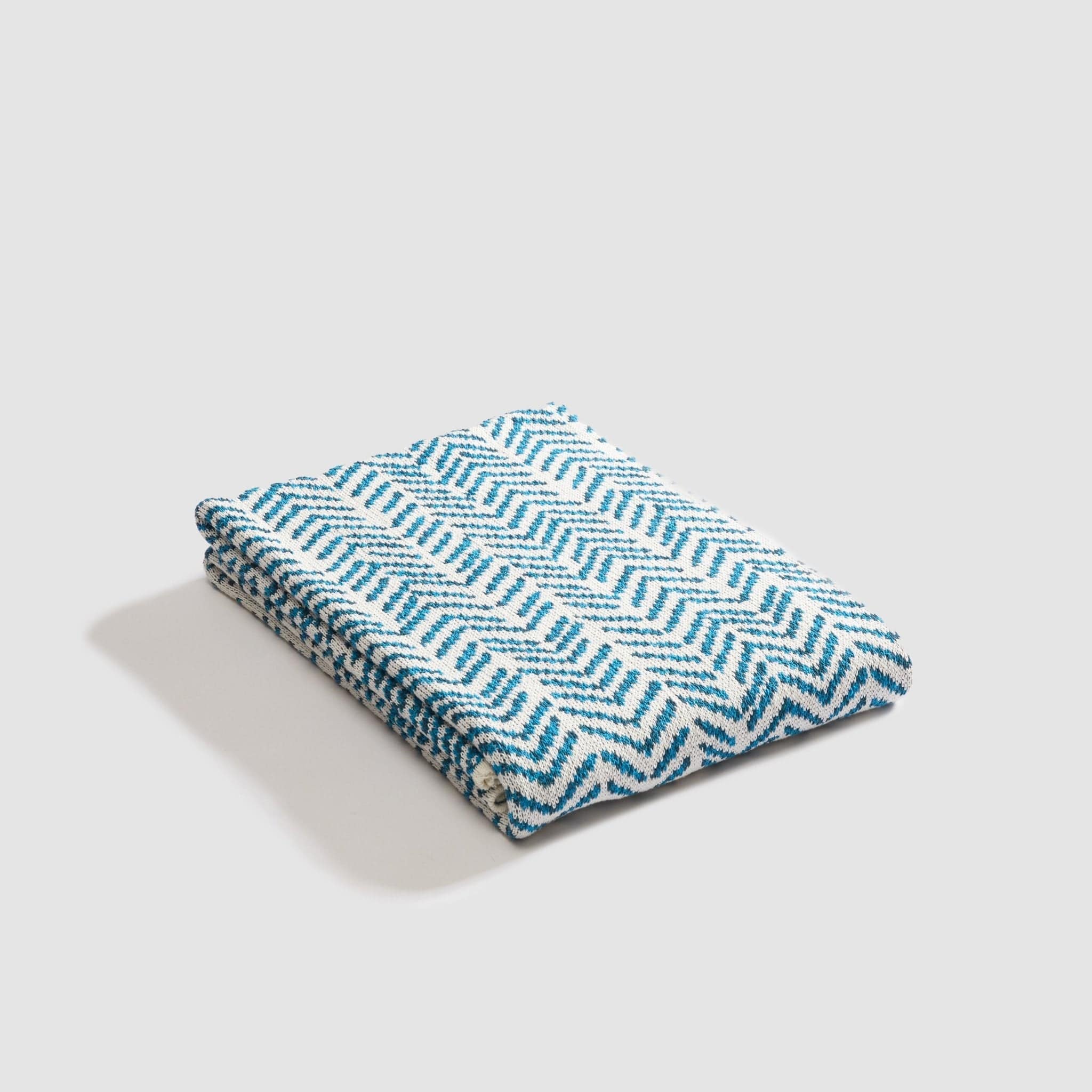 An In2Green Warrior Throw Blanket in teal, showcasing a bold blue and white zigzag pattern, lies folded against a plain backdrop, symbolizing strength and offering comfort.