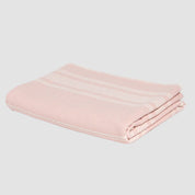 Against a white backdrop, the in2green Woven Twill Blanket, crafted from recycled cotton, displays its neatly folded design in light pink with thin white stripes.