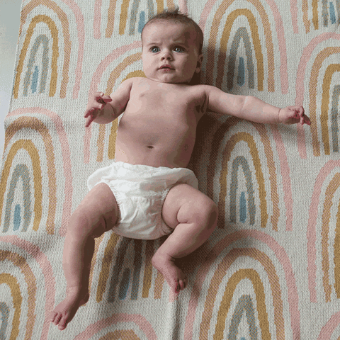A baby wearing a diaper lies on an Eco Throw, the Baby Rainbow Blanket crafted from recycled cotton by in2green.