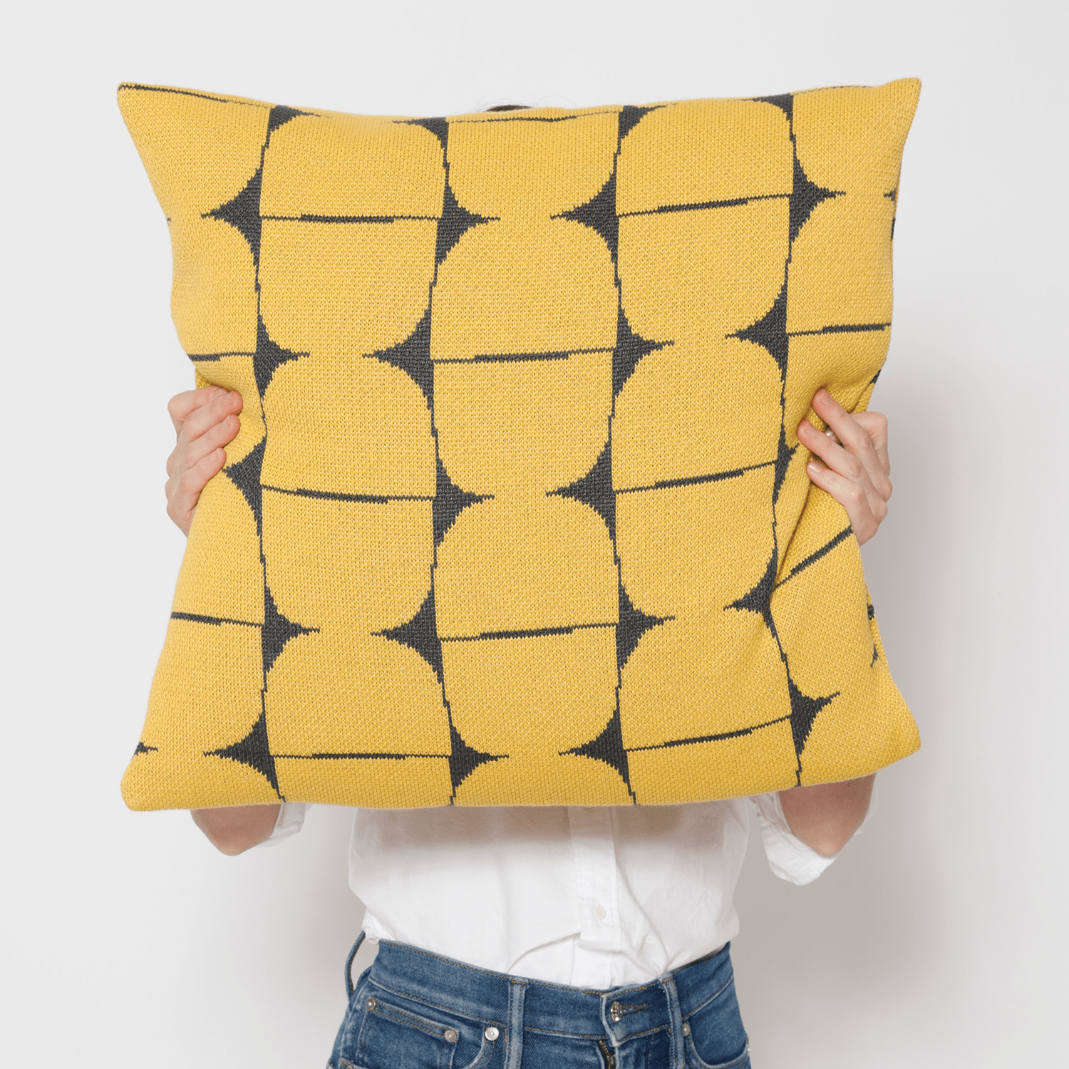 A person holds the Sale Poly Kaarna Pillow by Petra Kaksonen from in2green, featuring a bold black geometric pattern on its large yellow surface, partially obscuring their face. Dressed in a white shirt and jeans, they embody Nordic design aesthetics with chic simplicity.