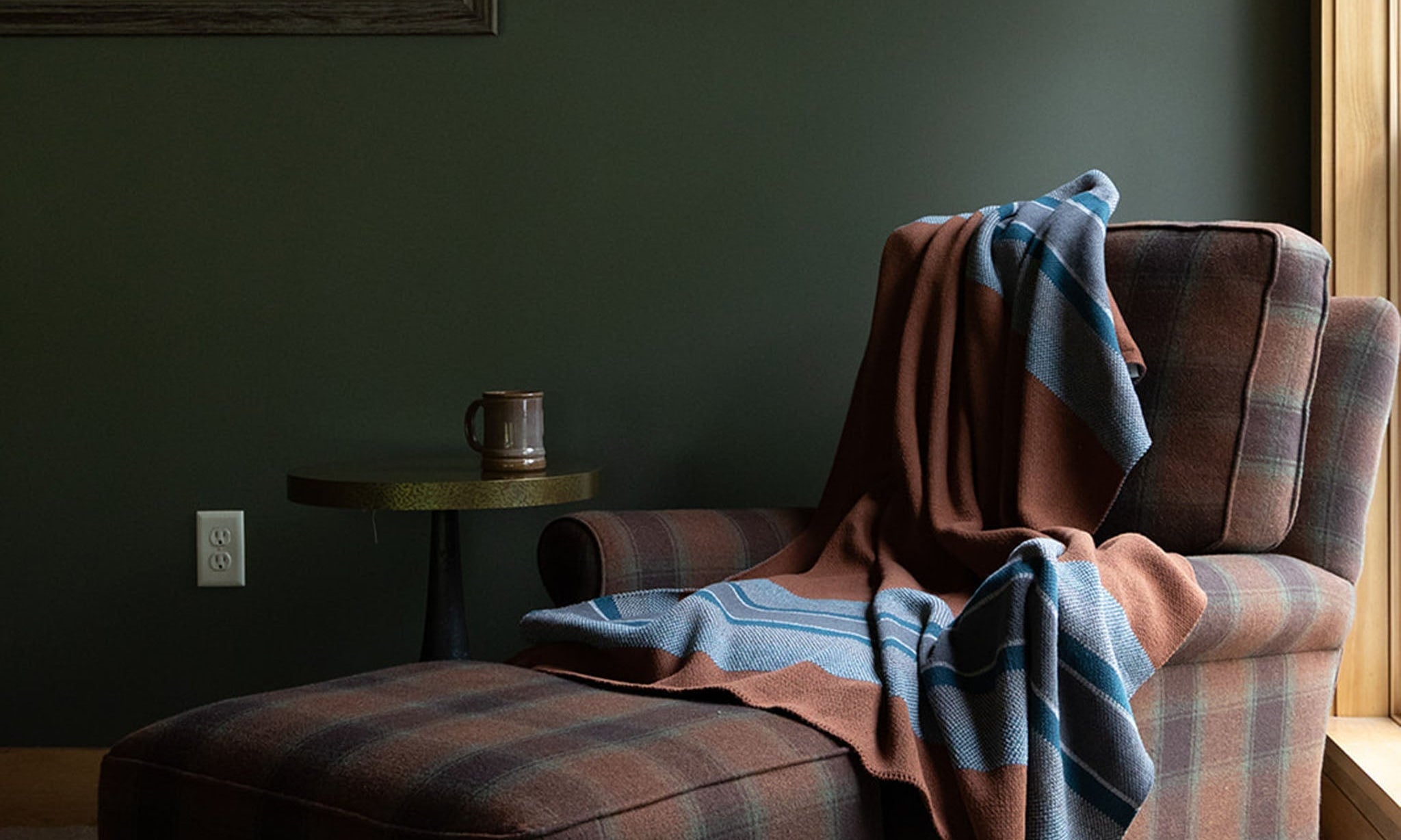 A brown blanket with blues stripes draped over the back of a brown plaid chair in a green room.