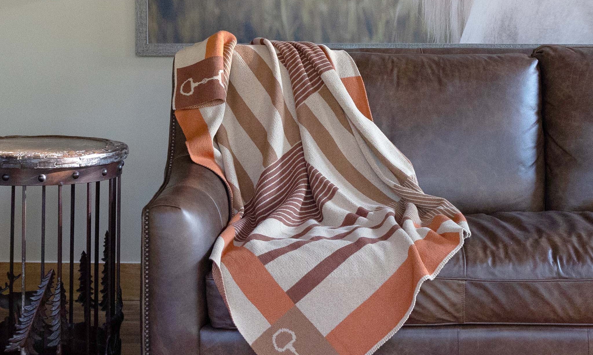 A striped orange and brown blanket with horse bits on a brown leather couch in a rustic living room.