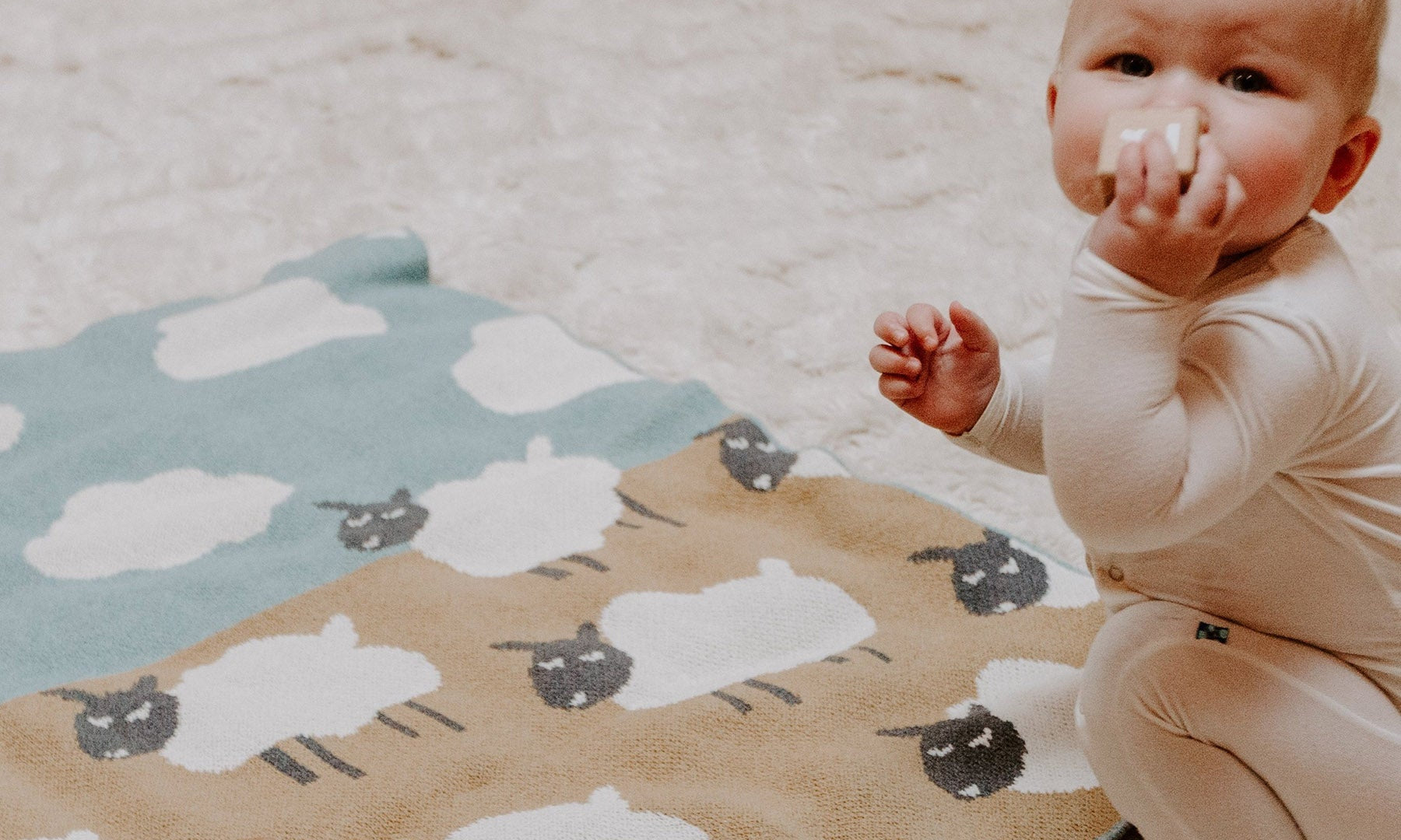 A baby in light-colored clothing sits on a soft carpet near a blanket with sheep patterns, holding a small white object and a wooden block with the letter "H.