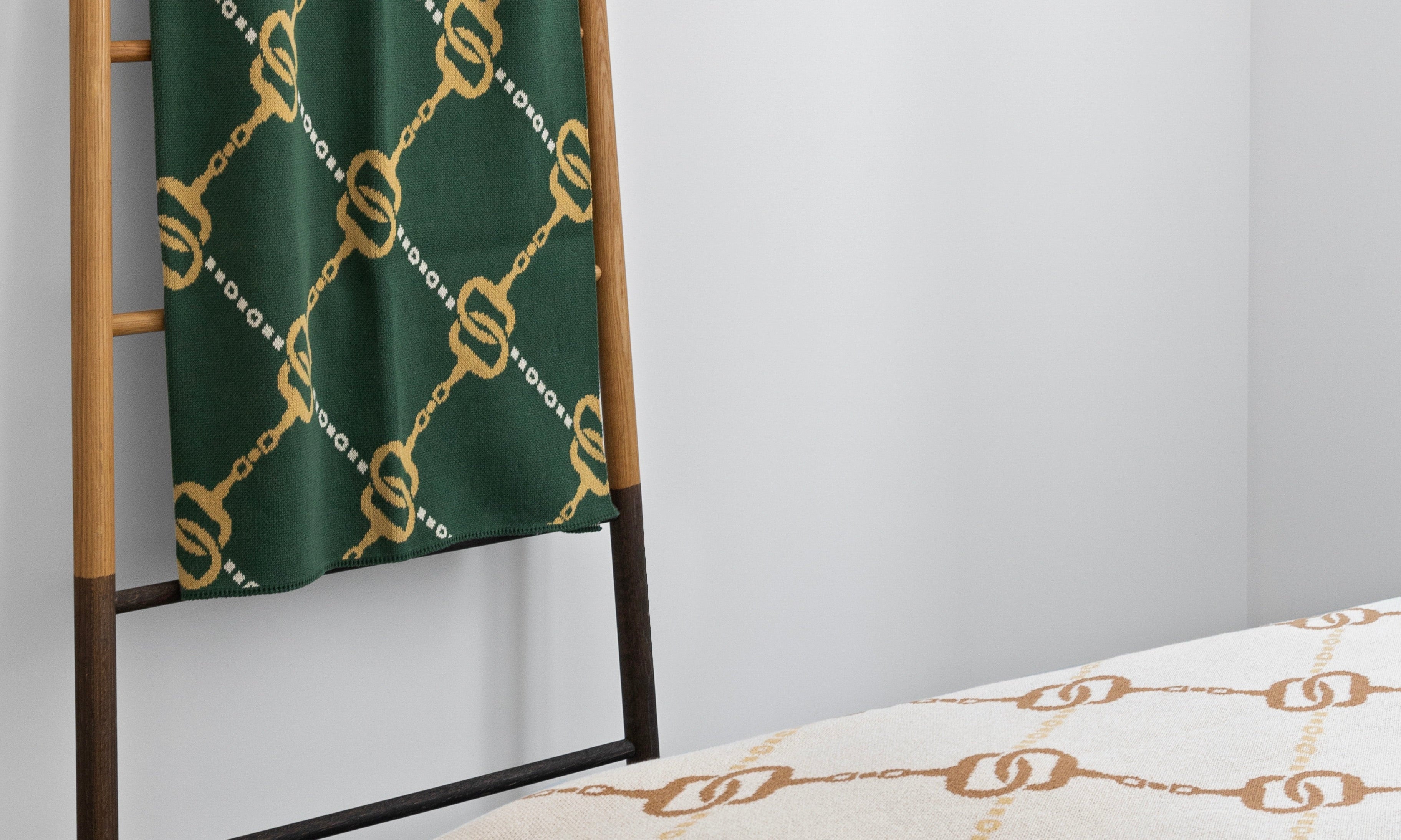 A green and gold patterned throw hangs on a wooden ladder next to a bed with a cream and gold chain motif blanket in a minimalistic room.