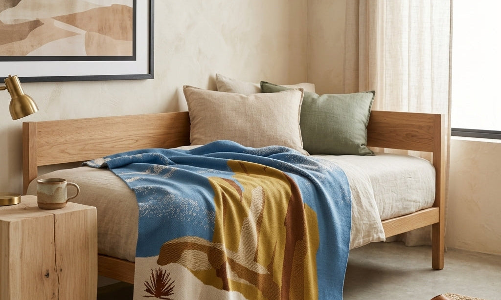 A green and gold patterned throw hangs on a wooden ladder next to a bed with a cream and gold chain motif blanket in a minimalistic room.
