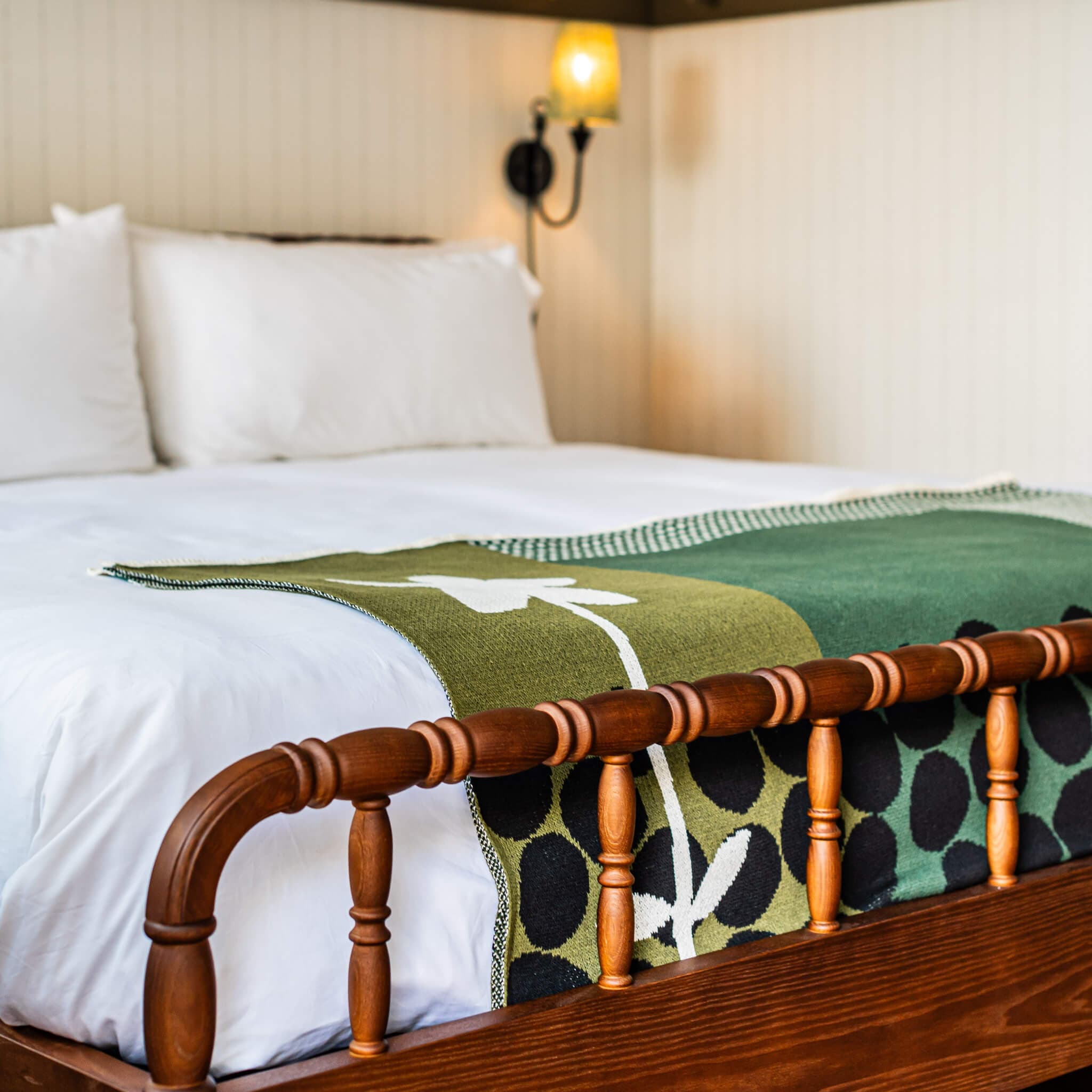 A softly lit room features a neatly made bed with white linens and pillows, a wooden footboard, and In2Green’s Rock Rose by Susy Pilgrim Waters—an eco-friendly green patterned blanket with hand-crafted appeal.