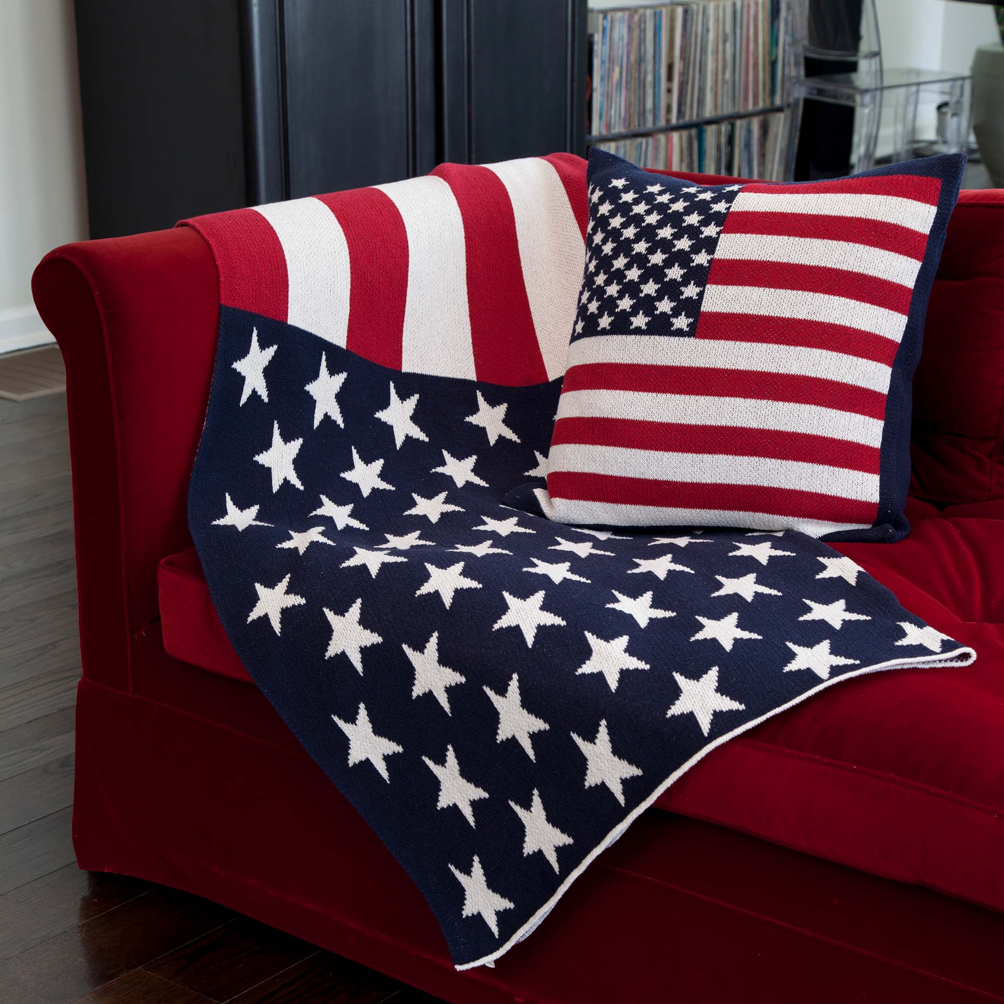 A red sofa is draped with the in2green Hometown Pride Flag – Cotton Sustainable Knit Blanket, featuring an American flag design, and is paired with a coordinating American flag pillow. Black shelves and a record collection are visible in the background.