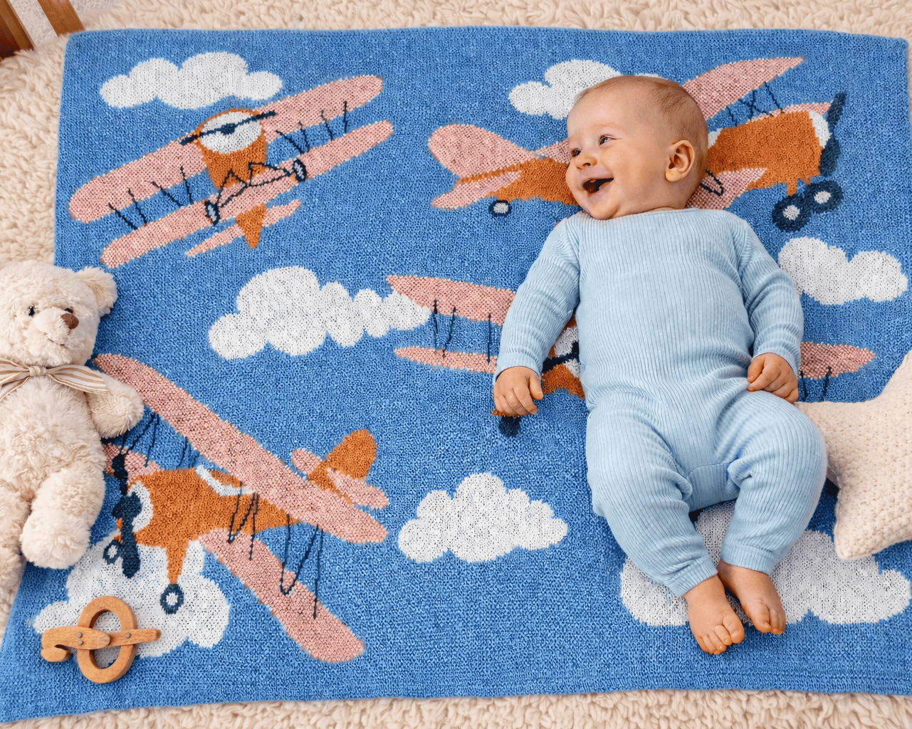 Baby lying on a blue blanket with airplane designs, surrounded by toys.