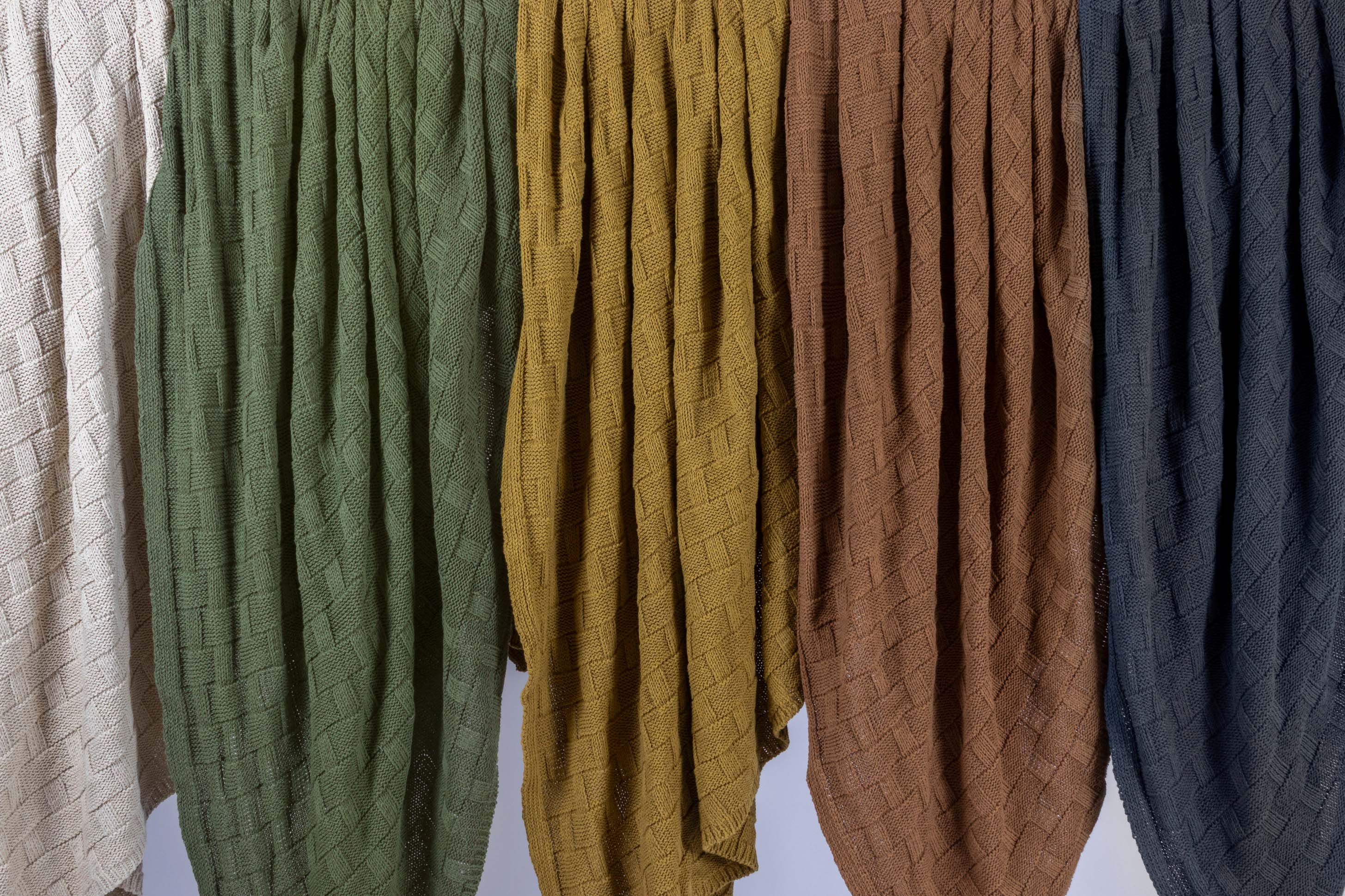 Five Basketweave Throw Blankets by in2green, made from recycled yarns in beige, green, yellow, brown, and dark blue, hang side by side against a light background.