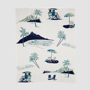 The Beach Toile Throw Blanket is USA-knit from recycled yarn and shows a tropical scene with palm trees, lounge chairs, lifeguard towers, and a mountain in blue and green on a white background.
