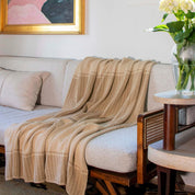 The in2green Block Canyon Throw Blanket is draped over a light-colored sofa with wooden armrests. A pillow, flowers on a side table, and framed artwork complete the cozy setting.