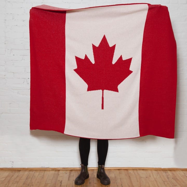 A person stands behind the in2green Hometown Pride Flag - Cotton Sustainable Knit Blanket, with only their lower legs and boots visible, in front of a white brick wall.