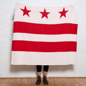 A person stands behind the in2green Hometown Pride Flag - Cotton Sustainable Knit Blanket, featuring the Washington, D.C. flag design with three red stars and two red bars on a white background; only their lower legs and feet are visible. This custom blanket is proudly made in the USA from recycled cotton.